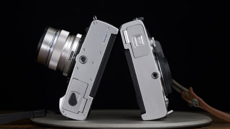 Two silver digital cameras stand on their sides, facing each other on a circular gray surface against a black background.
