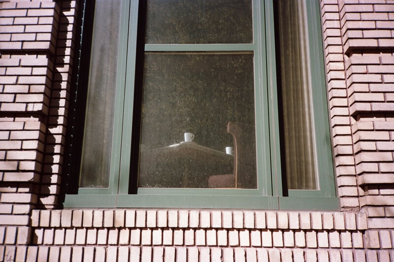 A view through a dusty window shows a table with two white mugs and a chair inside a brick building.