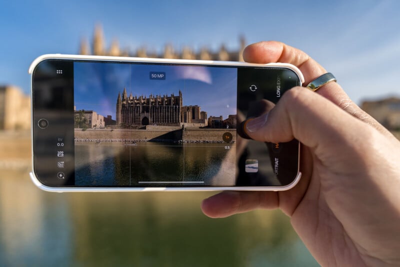 A hand holding a smartphone displays a camera interface showing a live view of a large stone cathedral reflected in a body of water.