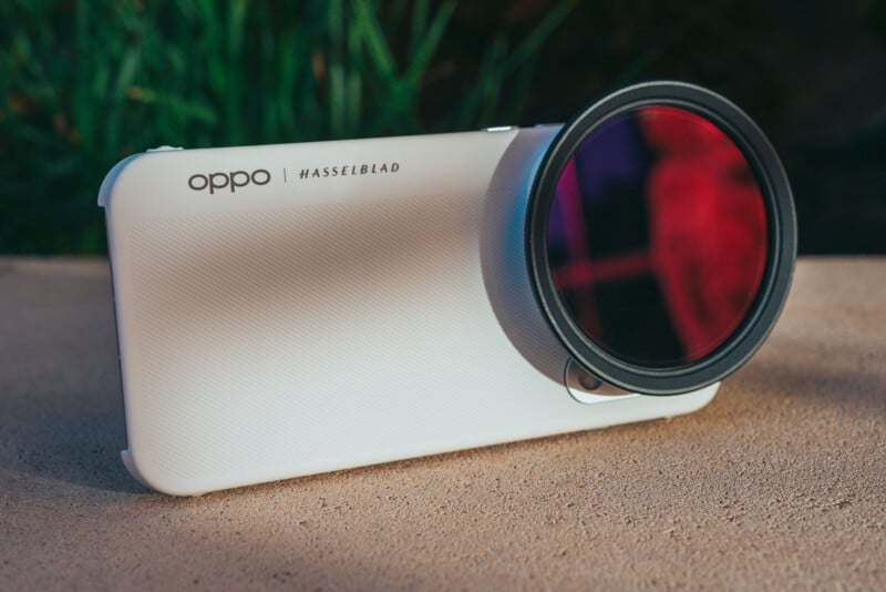 A white smartphone case with "oppo | HASSELBLAD" text is resting on a textured surface, featuring a large circular lens filter attached over the camera area.