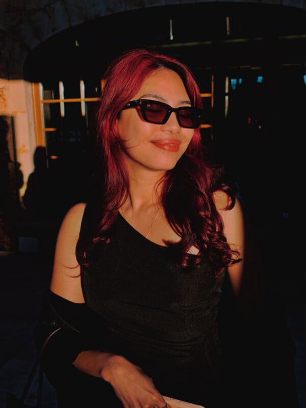 A person with vibrant red hair and sunglasses smiles while standing in warm, golden sunlight against a dark background.