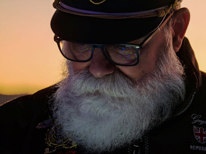 A close-up portrait of an older man with a thick white beard, wearing glasses and a captain's hat, looking downward against a sunset background.