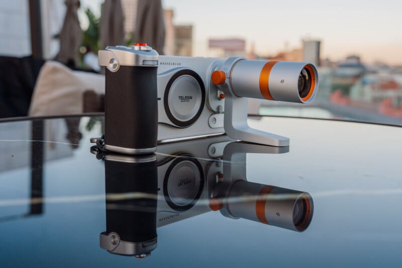 A silver and black camera with orange accents sits on a reflective black table, showing its reflection below.