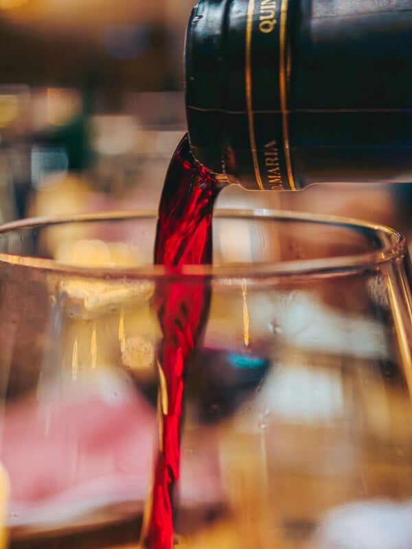 Red wine pours from a dark bottle labeled D. MARIA into a clear glass.