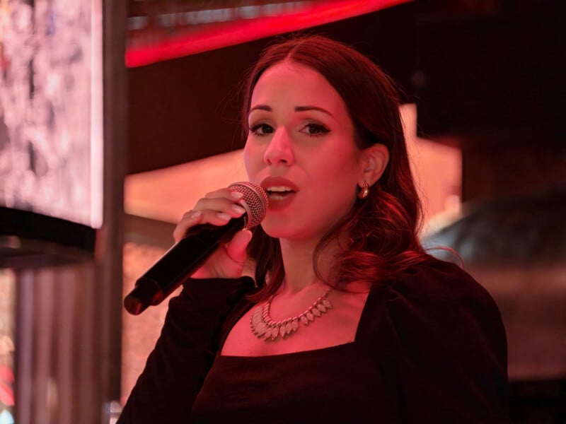 A woman with long brown hair wearing a black top and a beaded necklace speaks into a handheld microphone under red lighting.
