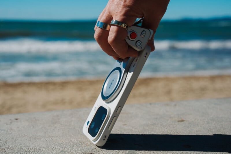 A hand wearing two rings holds a white smartphone case with a magnetic attachment and a small remote shutter button against a blurred beach background.