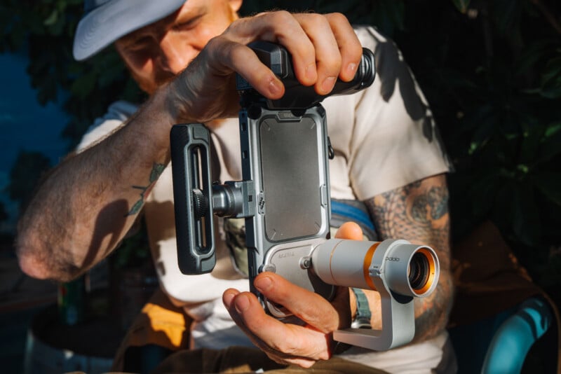 A person with tattooed arms holds a smartphone mounted in a specialized metal cage with an attached handle and a lens accessory.