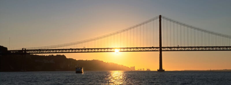 A suspension bridge spans across a body of water during a golden sunset, with a small boat traveling underneath.