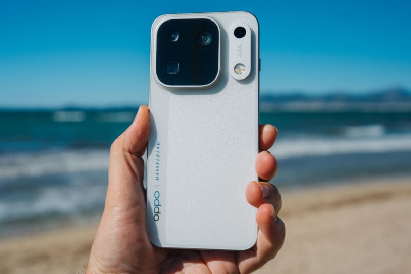 A hand holds a white smartphone with a large, black, rectangular camera module on the back against a blurred beach background.