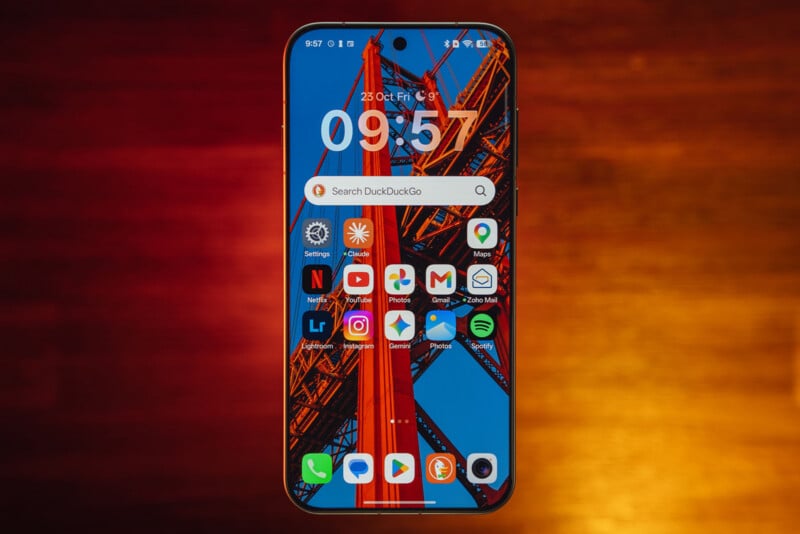A smartphone screen displays a home screen with a bridge wallpaper, a large digital clock, a DuckDuckGo search bar, and various app icons against a blurred red and orange background.