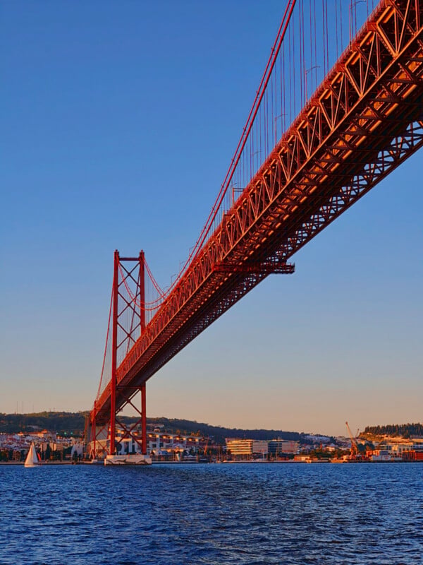 A low-angle view of a large red suspension bridge spanning across a body of water, with a small sailboat visible near the shore.