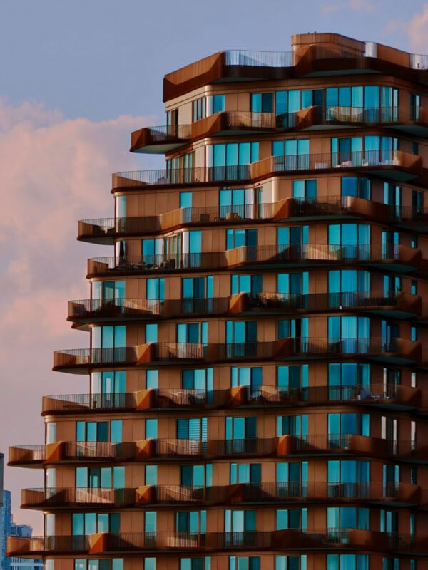 A modern, tiered apartment building with copper-toned metal cladding and glass balconies against a soft, pink-hued sky.