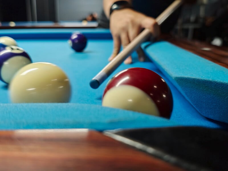 A person's hand holds a pool cue over a blue felt table, aiming toward a red and white striped billiard ball near the corner pocket.