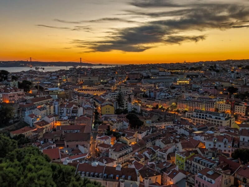 A high-angle view of a dense city with terracotta-roofed buildings at sunset, featuring a large suspension bridge in the distance.