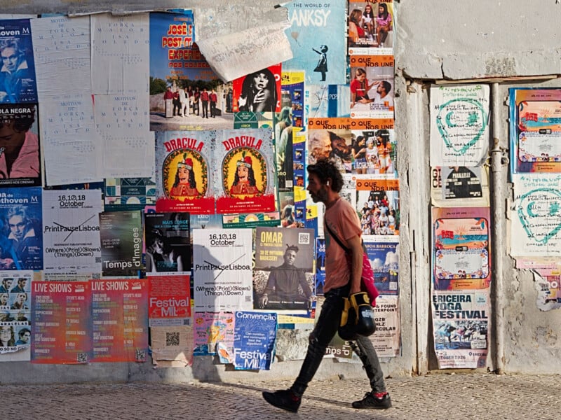 A man carrying boxing gloves walks past a wall covered in various event posters and flyers in an urban setting.