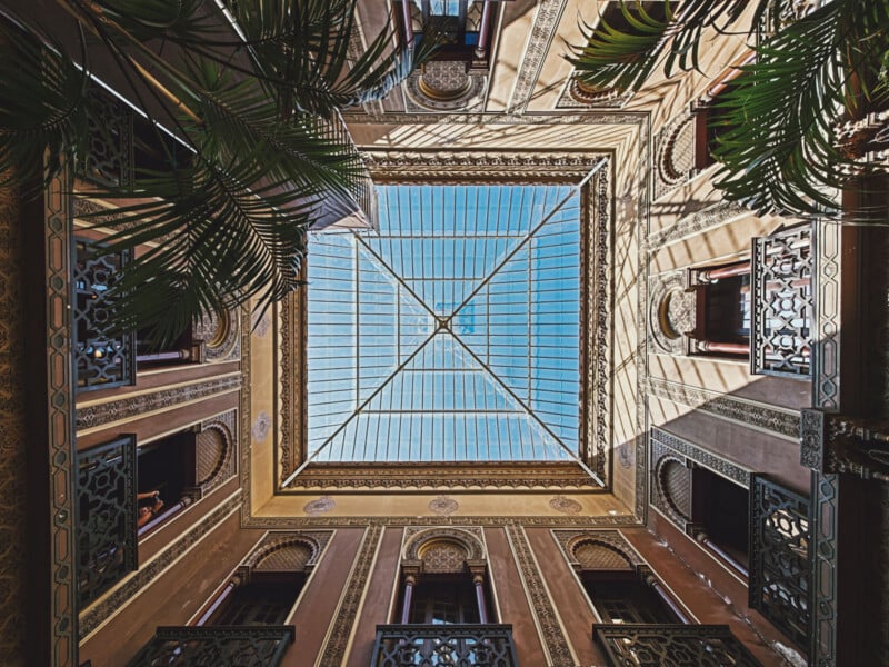 A low-angle view looking up through a glass skylight at the center of a multi-story building with ornate, Moorish-style architecture and palm fronds in the corners.