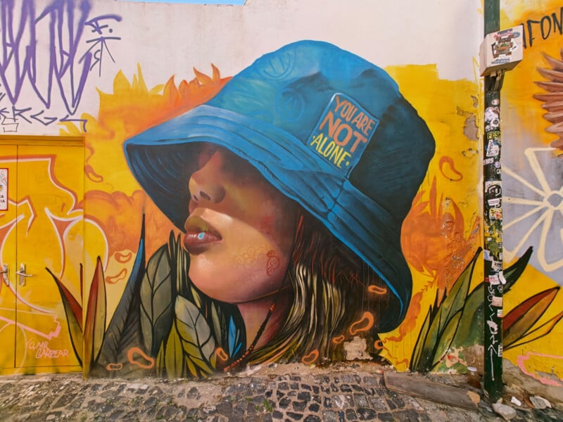 A vibrant mural on a textured wall depicts a person in profile wearing a large blue bucket hat with the text "YOU ARE NOT ALONE" printed on it, set against a yellow and orange background with stylized leaves.