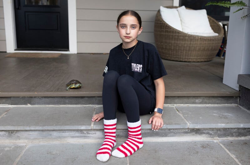 A young girl wearing a black t-shirt and red-and-white striped socks sits on stone steps next to a small turtle.