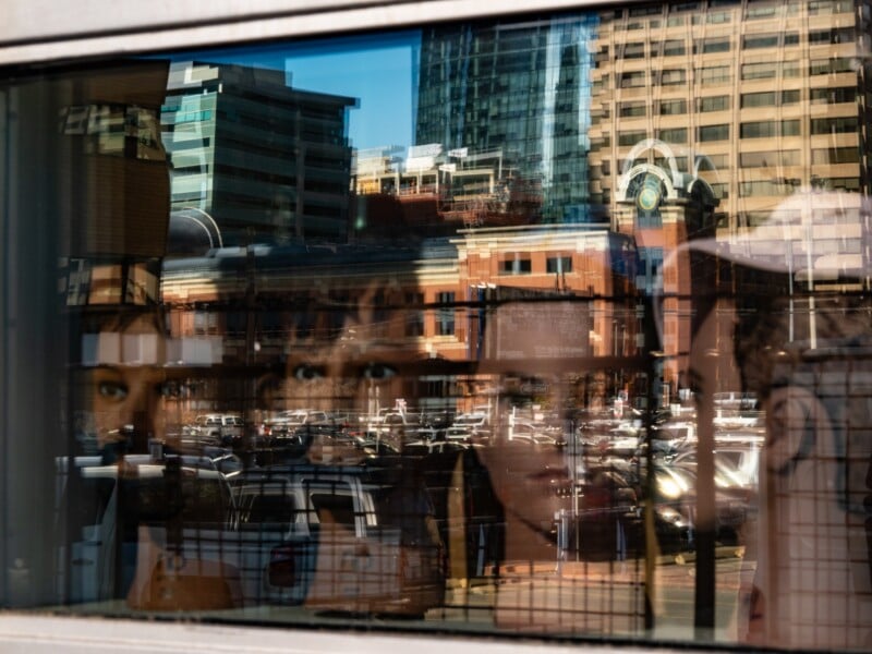Reflections of city buildings and a parking lot are superimposed over the faces of several mannequins displayed in a shop window.