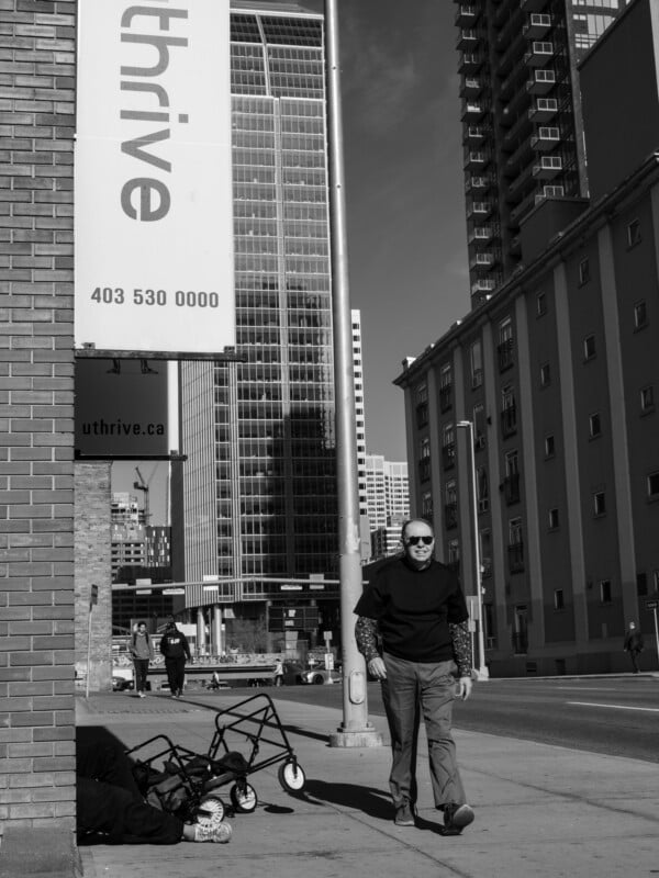 A man in sunglasses and a dark sweater walks down a city sidewalk past a brick building with a large "thrive" banner, while a person sits on the ground nearby with a small cart.