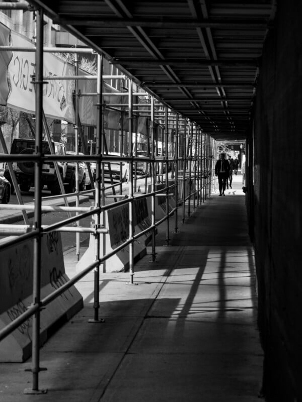 A man in a suit walks down a sidewalk protected by a metal scaffolding tunnel, with a street and parked vehicles visible to the left.