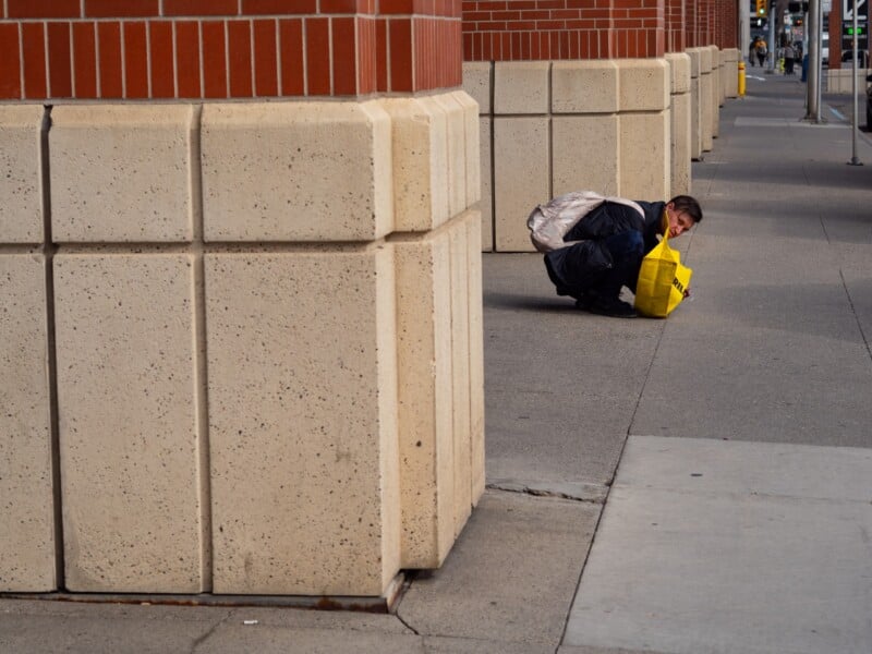 A person wearing a backpack crouches on a sidewalk next to a large brick pillar, looking down at a bright yellow bag on the ground.