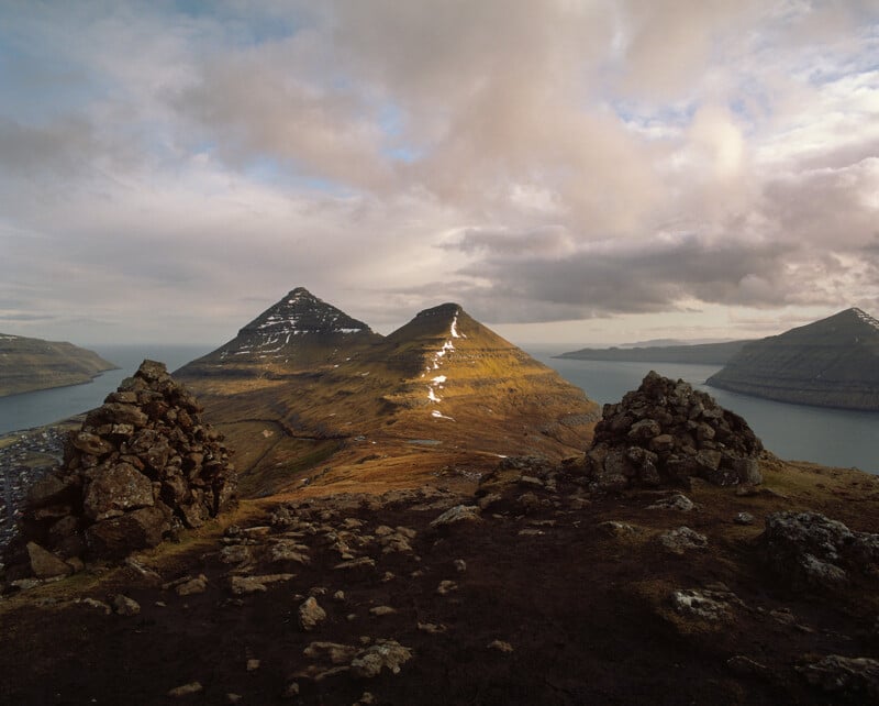 Two stone cairns sit on a dark, rocky foreground overlooking a valley with two prominent, grassy mountains and a bay.