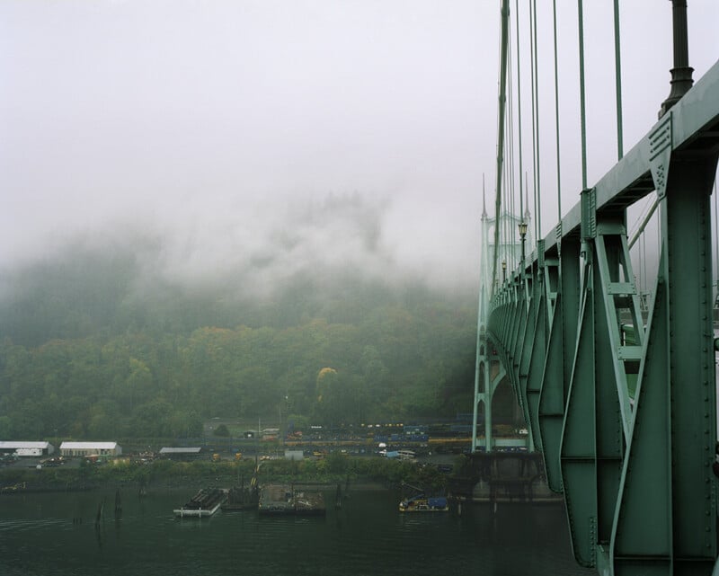 A large green steel suspension bridge spans across a river, with a foggy, forested hillside in the background.