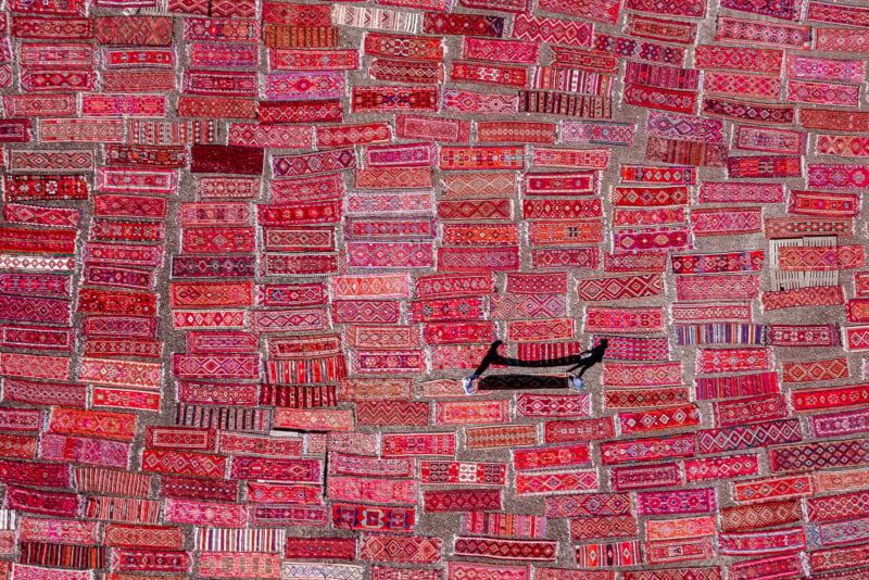 An aerial view shows many rectangular red patterned rugs laid out on the ground, with two people walking among them.
