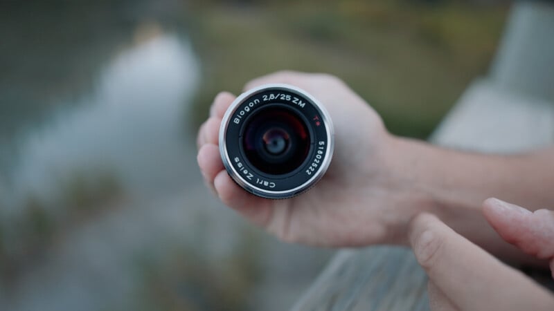 A person holds a silver Carl Zeiss Biogon 2.8/25 ZM camera lens in their hand against a blurred outdoor background.