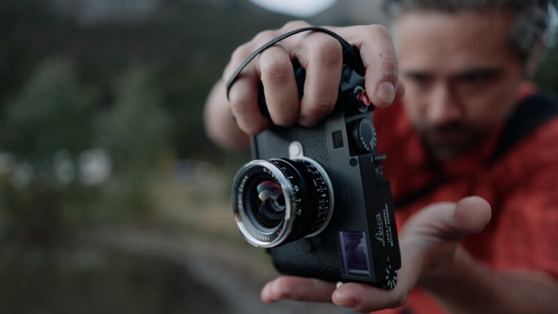 A person in a red shirt holds a black Leica camera with a Carl Zeiss Biogon lens toward the viewer.