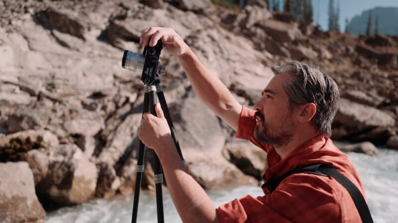 A man with graying hair and a beard, wearing a red shirt, adjusts a camera mounted on a tripod outdoors near a rocky stream.