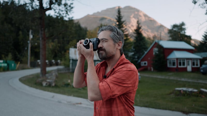 A middle-aged man with gray hair and a beard wearing a red shirt holds a camera to his eye to take a picture outdoors with a mountain in the background.