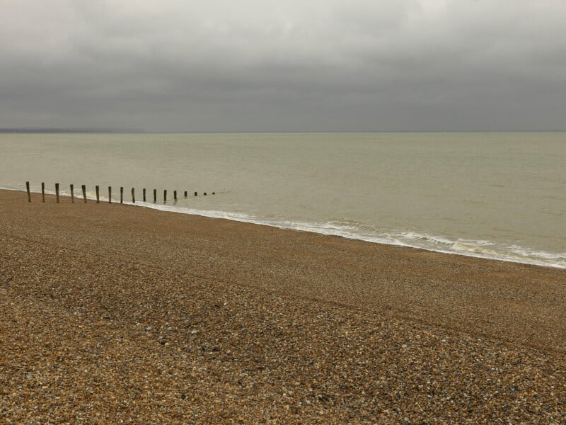 A row of wooden posts extends from a shingle beach into a calm, grey sea under a cloudy sky.