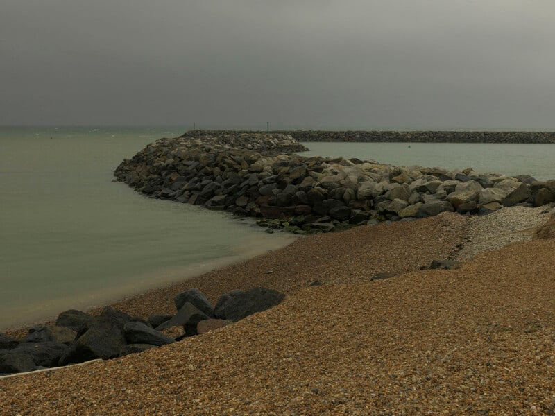 A curved stone breakwater extends into a calm, grey sea under an overcast sky, with a shingle beach in the foreground.