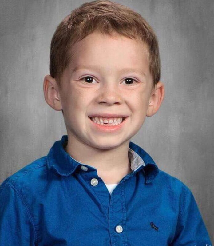 A young boy with light brown hair and a missing front tooth smiles at the camera while wearing a blue button-down shirt.