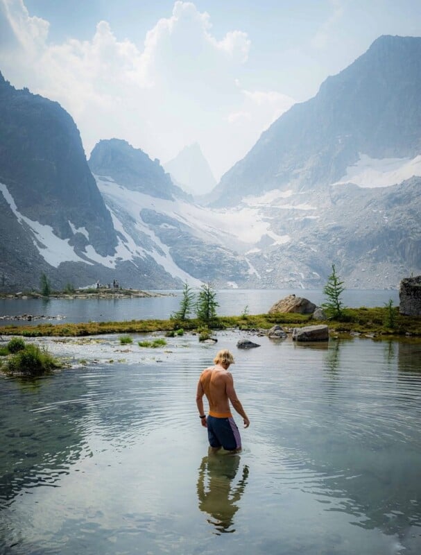 A shirtless man stands in the shallow, clear water of an alpine lake, facing away from the camera toward snow-capped mountains.