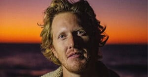 A close-up portrait of a man with wavy blonde hair and a light beard, illuminated by the orange glow of a sunset.