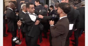 A reporter holding a DAZN microphone reaches out to adjust the tie of a man holding a camera on a red carpet.