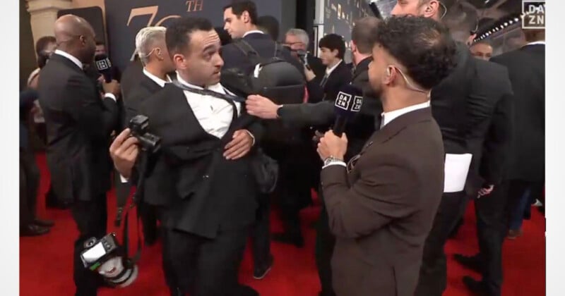 A reporter holding a DAZN microphone reaches out to adjust the tie of a man holding a camera on a red carpet.