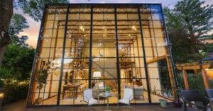 A modern, two-story house with a floor-to-ceiling glass grid facade overlooks a wooden deck with outdoor seating at dusk.