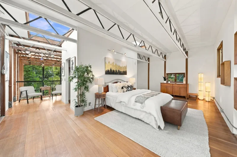 A spacious, open-plan bedroom with high ceilings, exposed trusses, and a skylight, featuring a bed, a leather bench, wooden furniture, and an adjacent sunroom with large windows.