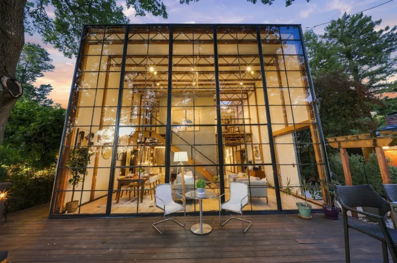 A two-story glass-walled house stands on a wooden deck at dusk, featuring a dining area and living room visible through the windows with two chairs and a small table in the foreground.