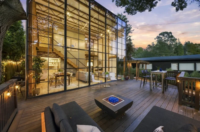 A modern house with a floor-to-ceiling glass wall overlooks a wooden deck featuring a fire pit, outdoor seating, and string lights at dusk.