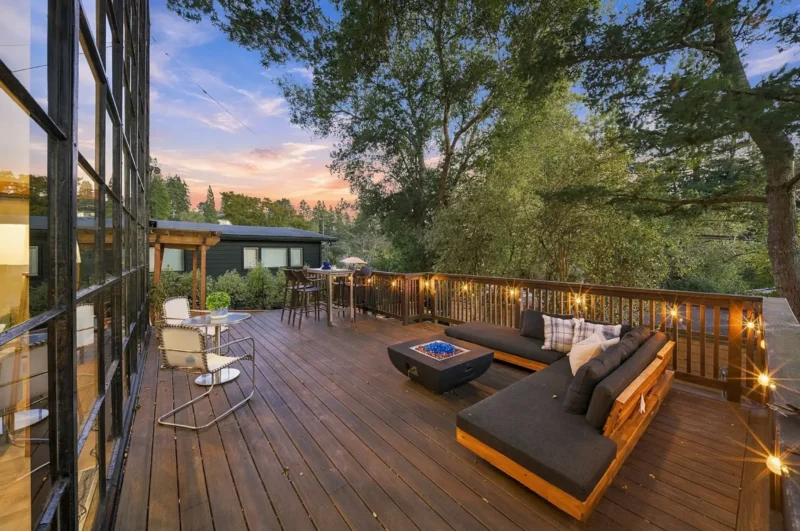 A wooden deck at dusk features a large L-shaped outdoor sofa with a fire pit, a small bistro table with two chairs, and a high-top table, all illuminated by string lights along the railing.
