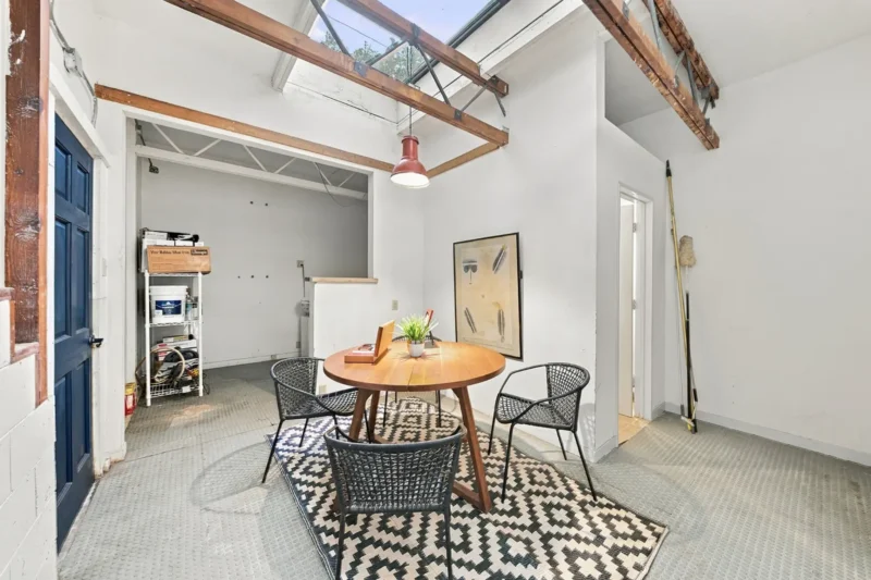 A dining area with a round wooden table and three black woven chairs sits on a patterned rug under a skylight with exposed wooden beams.