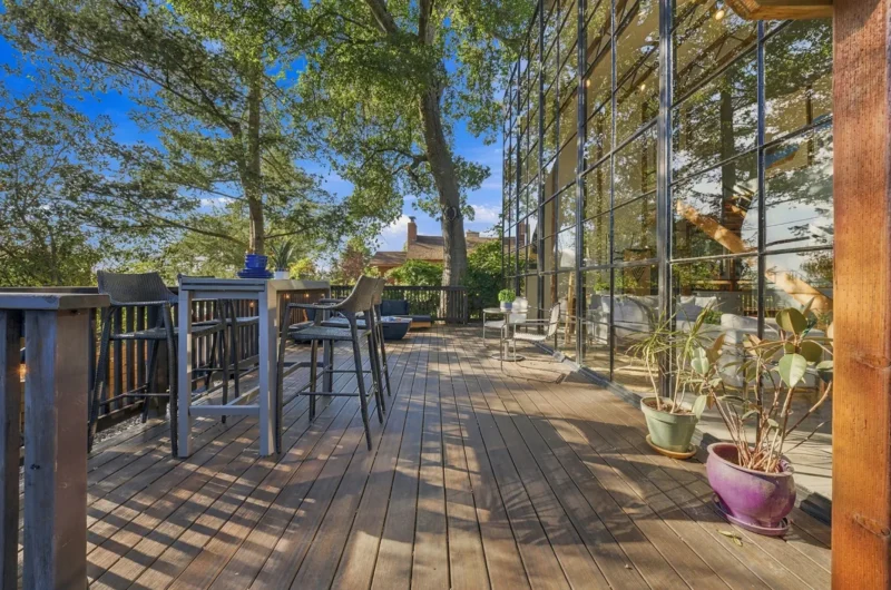 A wooden deck features a tall outdoor dining table with four chairs, a small bistro set, and potted plants, all adjacent to a large glass-walled building under a clear blue sky.