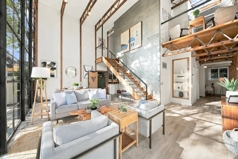A bright, open-concept living room features high ceilings, a wooden staircase, a gray sofa, two armchairs, and large windows.