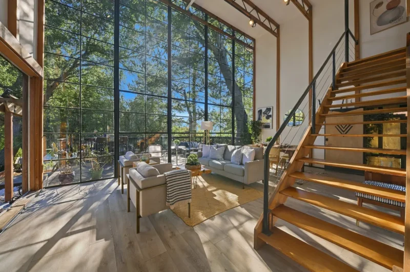 A sunlit living room features floor-to-ceiling windows, a gray sofa, two armchairs, a wooden staircase, and light wood flooring.