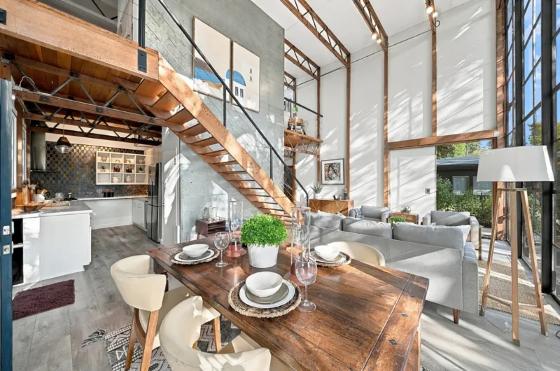 A bright, open-plan living and dining area features a wooden dining table in the foreground, a staircase leading to a loft, a modern kitchen, and a living room with a grey sofa under high ceilings.